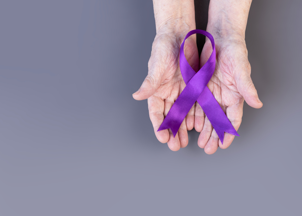 Hands of elderly woman hold purple ribbon on gray background. Alzheimer's awareness concept.